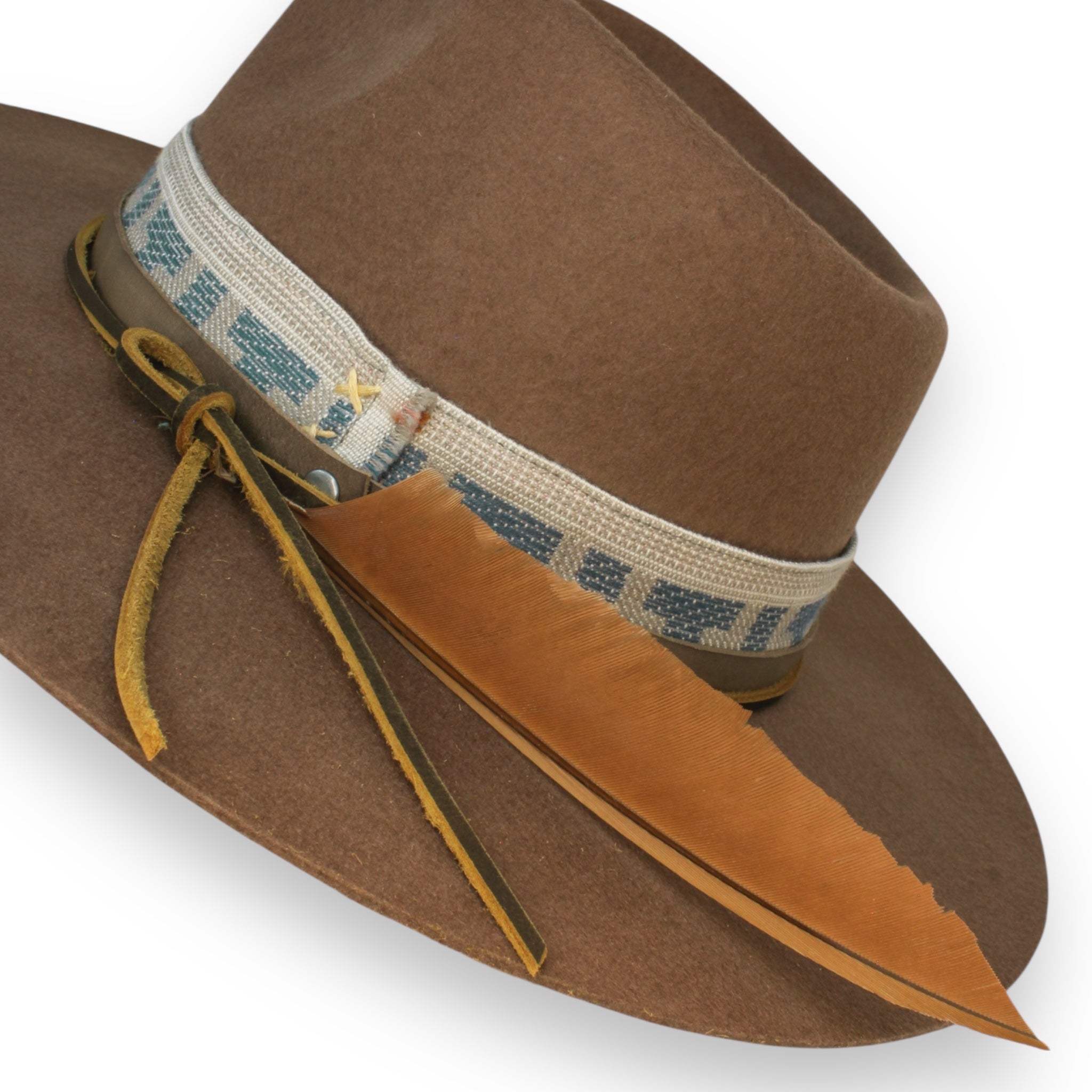 A detail shot of a brown fedora with a wide brim. The hat has a textile band, light brown leather band, looped leather hat tie, and a large feather.