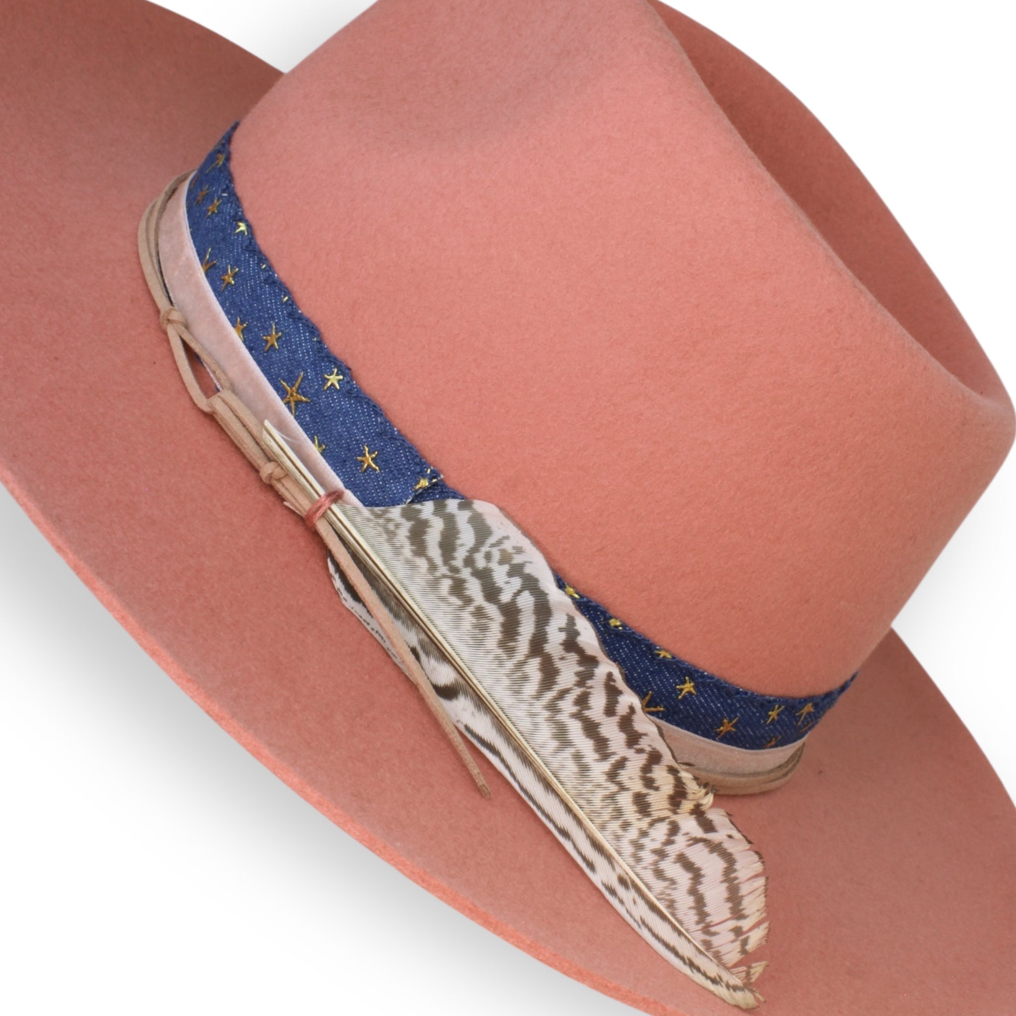 A detailed view of a felt coral colored hat with blue ribbon and a large white and brown feather.