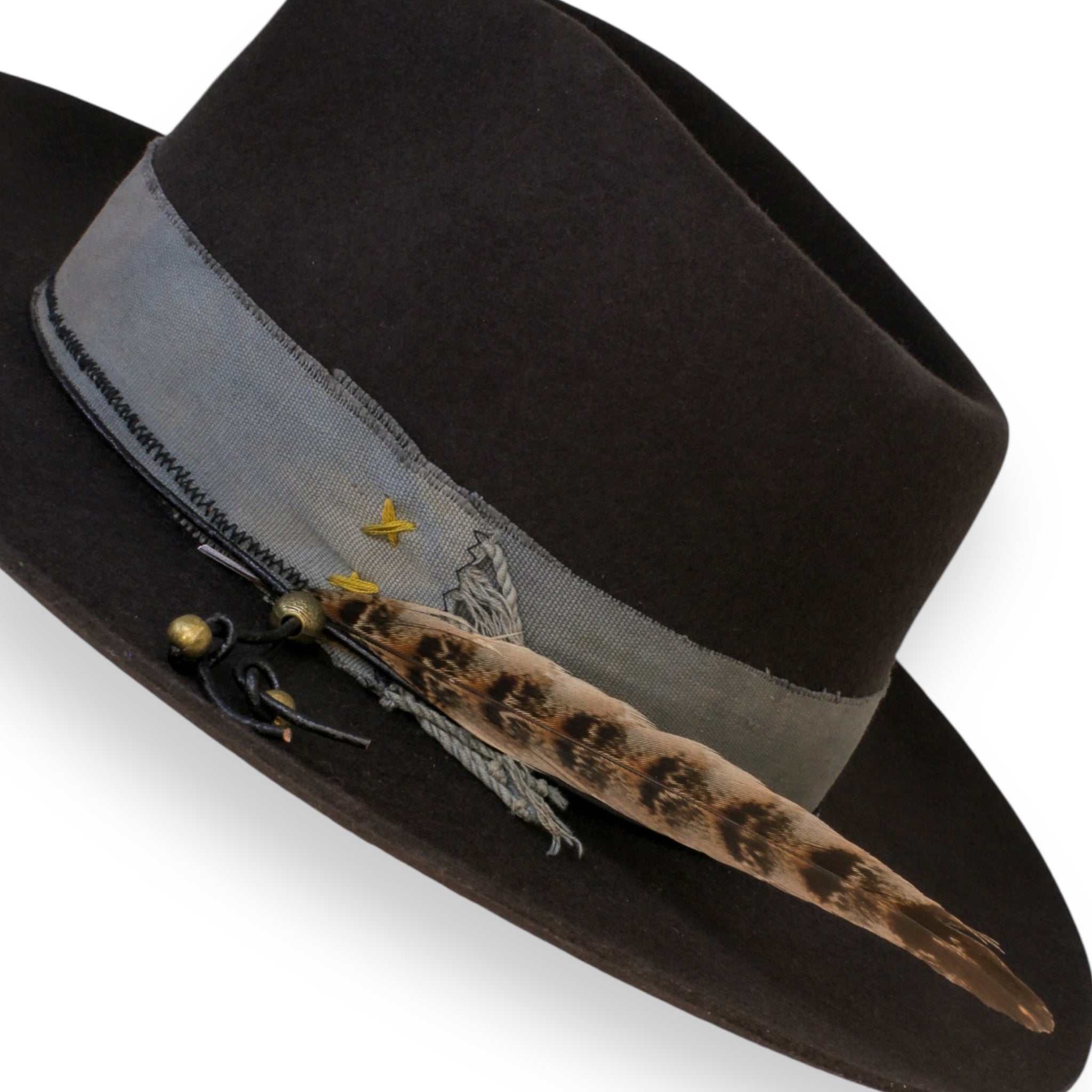 A detailed shot of the felt fedora. The textile band has dark muted yellow x stitches. The feather is long and slim. Three brass beads are on the ends of the hat tie.