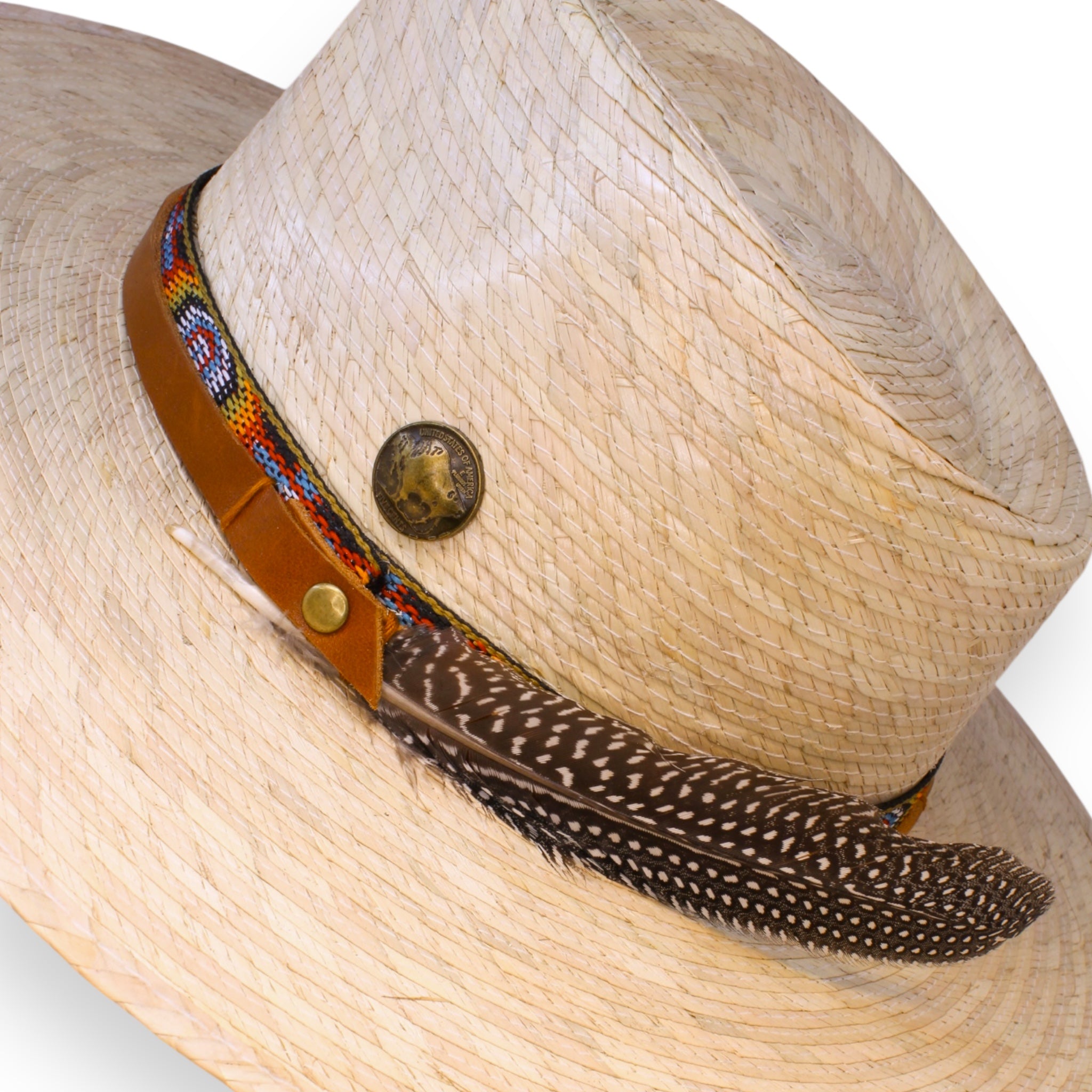 The natural palm hat from a closeup view showing off the intricacies in the native textile, tan leather band, brass buffalo coin, and the large spotted feather.