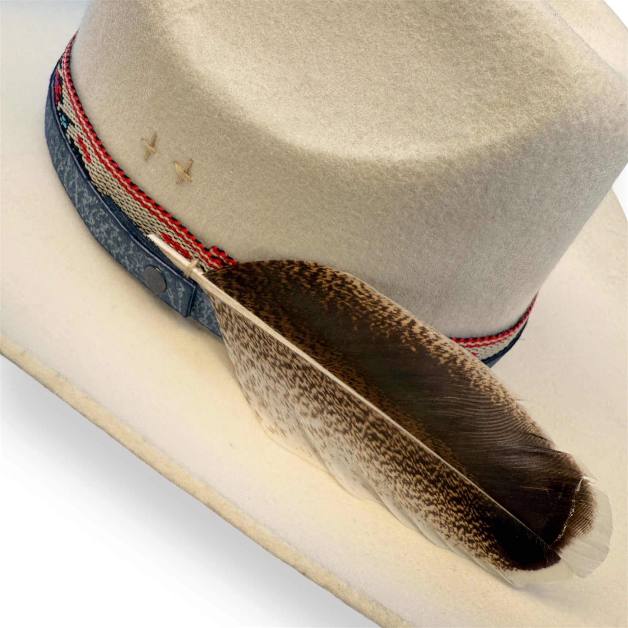 Detailed view of the off-white cowboy hat with a dark blue leather hat band, indigenous patterned ribbon, embroidered crosses, and a large feather.