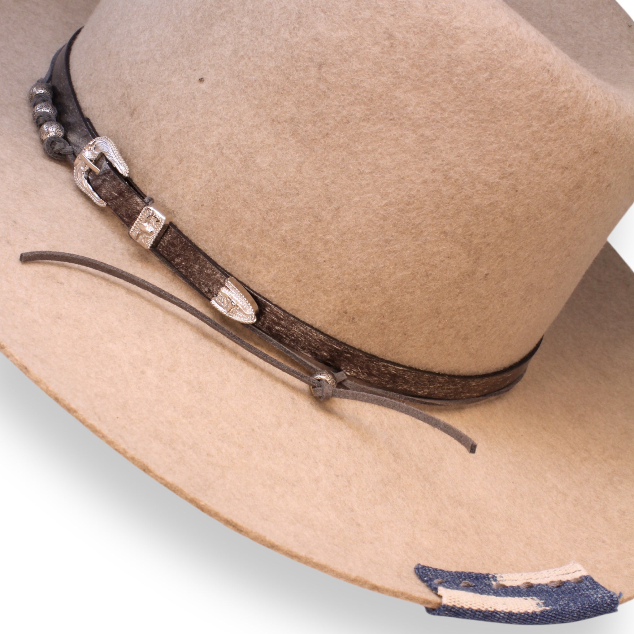 A detailed view of the speckled oatmeal colored fedora featuring a leather hat band with silver hardware, silver beads, and a blue and white textile patch.