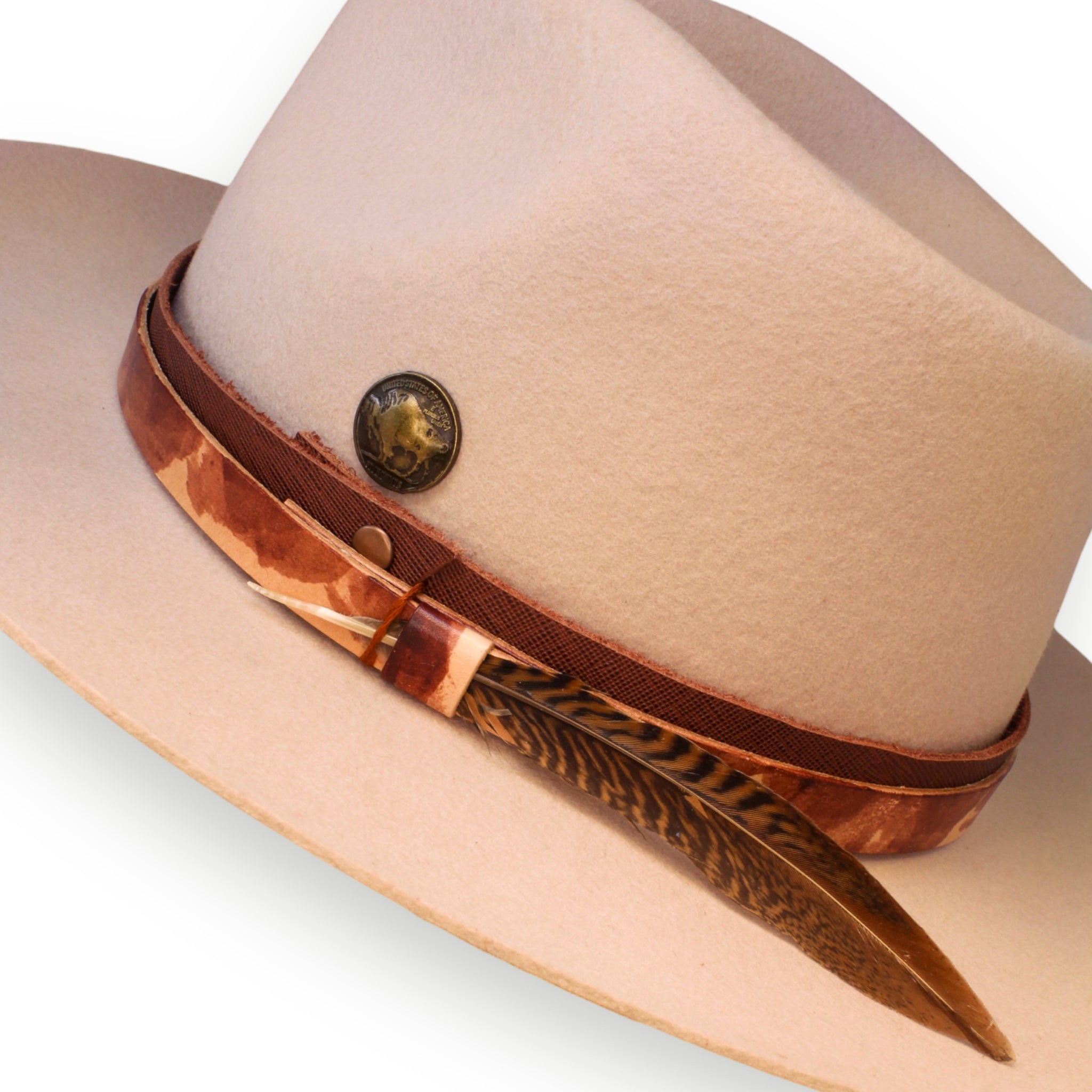 A detailed shot of an off-white felt hat. On the hat is a red leather band, a multicolored hat band, a bronze buffalo coin, and a red and black striped feather.